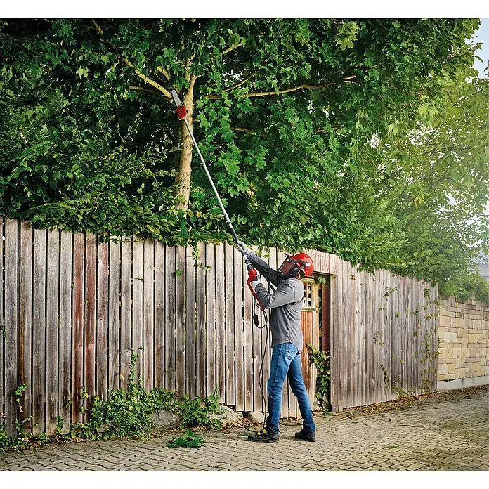 Einhell Cortasetos eléctrico GC-HC 90/2046 T - 900 W - Un hombre está usando una motosierra de pértiga para cortar ramas de un árbol. La motosierra es roja y plateada.
