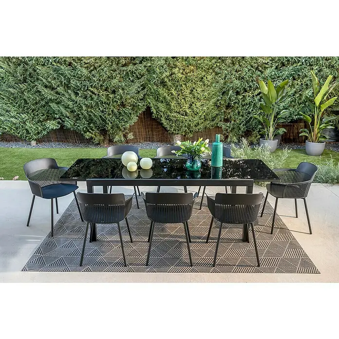 Mesa de jardín extensible Paris - L x An x Al: 180 x 90 x 75 cm, Aluminio/Vidrio, Negro - Una moderna mesa de jardín negra con sillas a juego sobre una alfombra geométrica, rodeada de exuberante vegetación.
