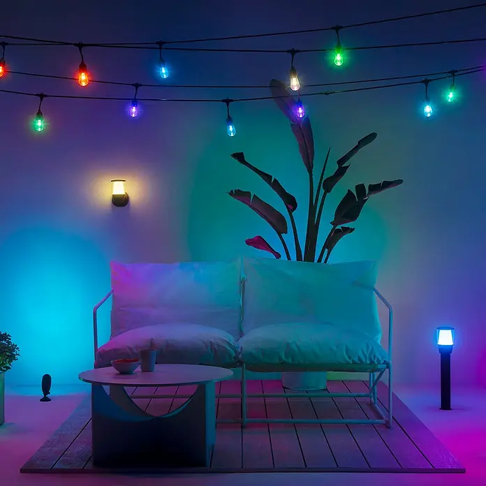 WiZ Außensockelleuchte Elpas Erweiterung - 4 W, RGBW, 10,85 x 10,85 x 51,8 cm, Mit Leuchtmittel - Bunte Lichterkette illuminiert eine gemütliche Terrasse mit weißen Möbeln und Pflanzen. Die Lichter sorgen für eine stimmungsvolle Atmosphäre.