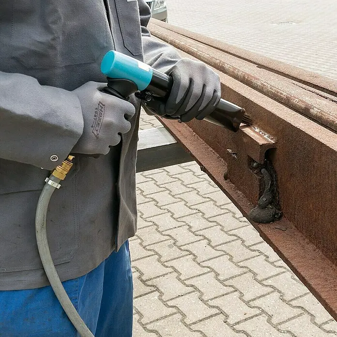 Hazet Nadelentroster 9035-5 - Betriebsdruck: 6,3 bar, Luftverbrauch: 170 l/min - Ein Arbeiter verwendet ein Nadelpistole, um Rost von einem Stahlträger zu entfernen. Die Pistole ist blau und schwarz mit einem grauen Schlauch.
