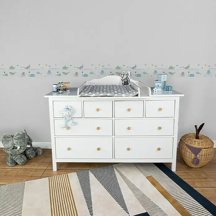 A.S. Création Lovely Kids Tapetenborte - Kunststoff, Wale - Weißer Wickeltisch mit neun Schubladen und runden Holzknöpfen in einem Kinderzimmer mit Spielzeug und Teppich.
