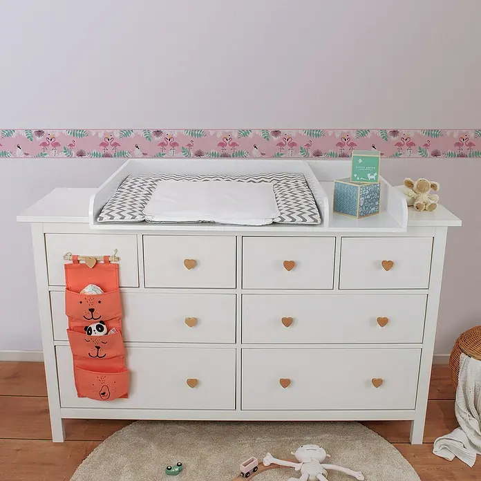 A.S. Création Lovely Kids Tapetenborte - Kunststoff, Flamingos - Eine weiße Wickelkommode mit Herzgriffen und einer orangefarbenen Aufbewahrungstasche mit Tiergesichtern für Windeln und Babypflegeprodukte.