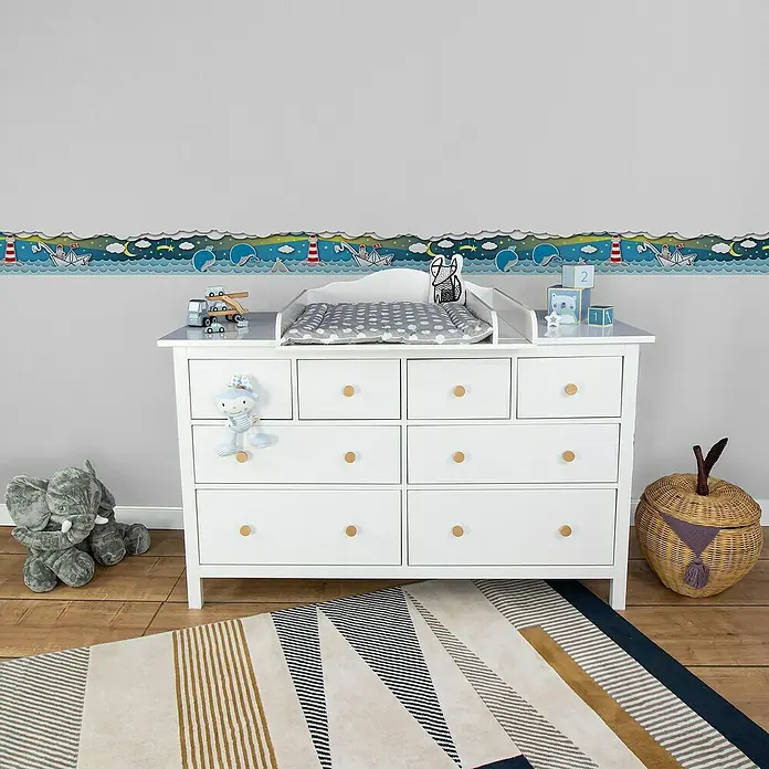 A.S. Création Lovely Kids Tapetenborte - Kunststoff, Wale mit Sternen - Ein weißer Wickeltisch mit bunten Motiven an der Wand, Stofftier und Spielzeugblöcken, ideal für ein Kinderzimmer.