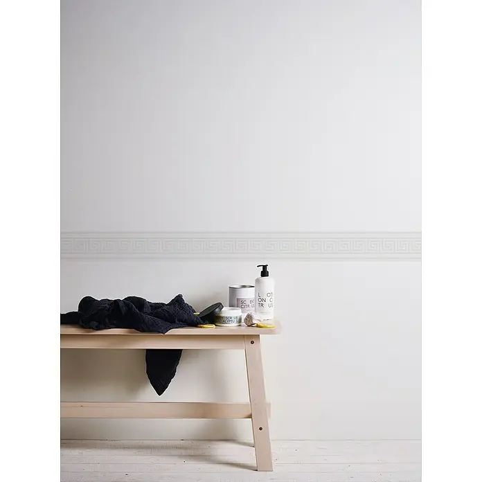 A.S. Création Only Borders 11 Tapetenborte Mäander - Weiß, 5 x 0,13 m, Selbstklebefolie - Auf einem hellen Holztisch arrangiert sind eine dunkle Stoffserviette, verschiedene Kosmetikprodukte und Zitronenscheiben vor einer weißen Wand mit Zierleiste.