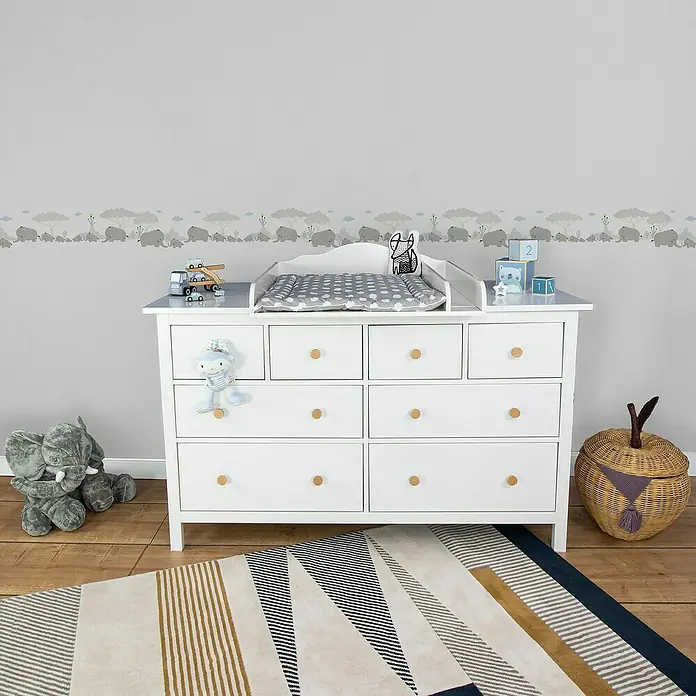 A.S. Création Only Borders 11 Tapetenborte Elefanten - Kunststoff, Grau/Weiß - Eine weiße Wickelkommode mit Holzgriffen, mit Spielzeug dekoriert, steht auf einem gemusterten Teppich in einem Kinderzimmer.