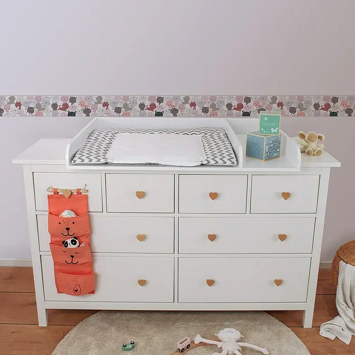 A.S. Création Lovely Kids Tapetenborte - Kunststoff, Kätzchen Rosa - Eine weiße Kommode mit einer Wickelauflage, Stoffwindeltasche und herzförmigen Griffen, die ein Kinderzimmerambiente ausstrahlt.