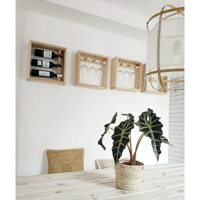 Astigarraga Botellero Píxel - L x An x Al: 12 x 39,6 x 40 cm, Madera, 3 botellas - Estantes de pared de madera clara que exhiben botellas de vino y copas sobre una planta de interior en maceta con cesta de mimbre.