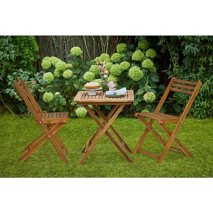 Sunfun Balkontisch - L x B x H: 65 x 65 x 74 cm, Akazie, Klappbar, Quadratisch, Natur - Einladende Gartenszene mit einem Holztisch und zwei Stühlen, gedeckt mit Kuchen, Blumen und Geschirr, umgeben von üppigem Grün.