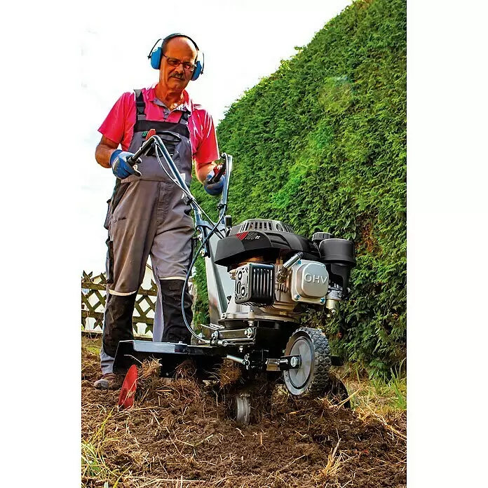 Scheppach Benzin-Bodenhacke MTP560 - 2.700 W, 4-Takt - Ein Mann bedient eine silberne und schwarze Gartenfräse mit '150cc OHV' Motor, die braune Erde neben einer grünen Hecke im Garten umgräbt.