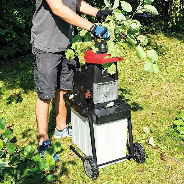 Scheppach Elektro-Leisehäcksler GS60 - Max. Aststärke: 4,5 cm, 2.800 W - Mann füttert grüne Äste in einen schwarz-roten Holzhäcksler mit weißem Auffangbehälter im Garten.