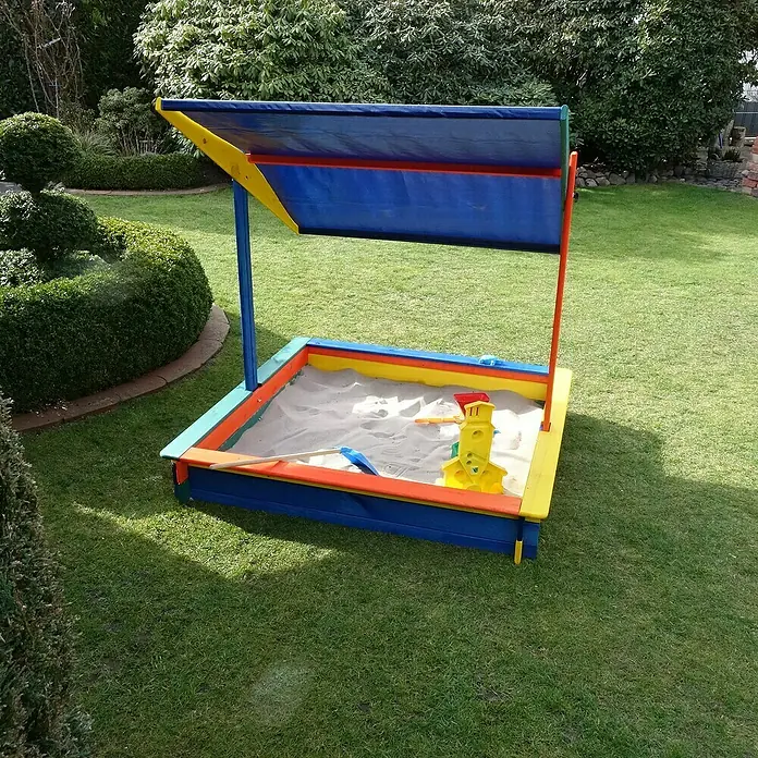 Promadino Sandkasten Nadine140 x 140 x 13 cm, Kiefer, Höhenverstellbares Dach - Ein farbenfroher Sandkasten mit Sonnenschutz, gefüllt mit Sand und Spielzeug, steht auf einem gepflegten Rasen im Garten.