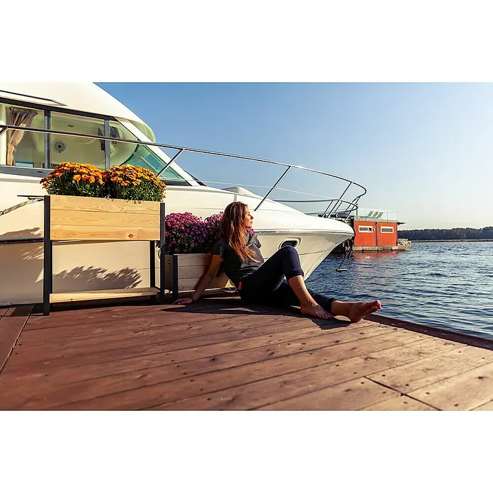 Upyard Hochbeet Modernbox hoch - 78 x 36 x 72 cm, Holz, Natur/Schwarz - Eine Frau entspannt auf einem Holzsteg neben einem eleganten Boot, das mit blühenden Blumenkästen geschmückt ist, an einem sonnigen Tag.