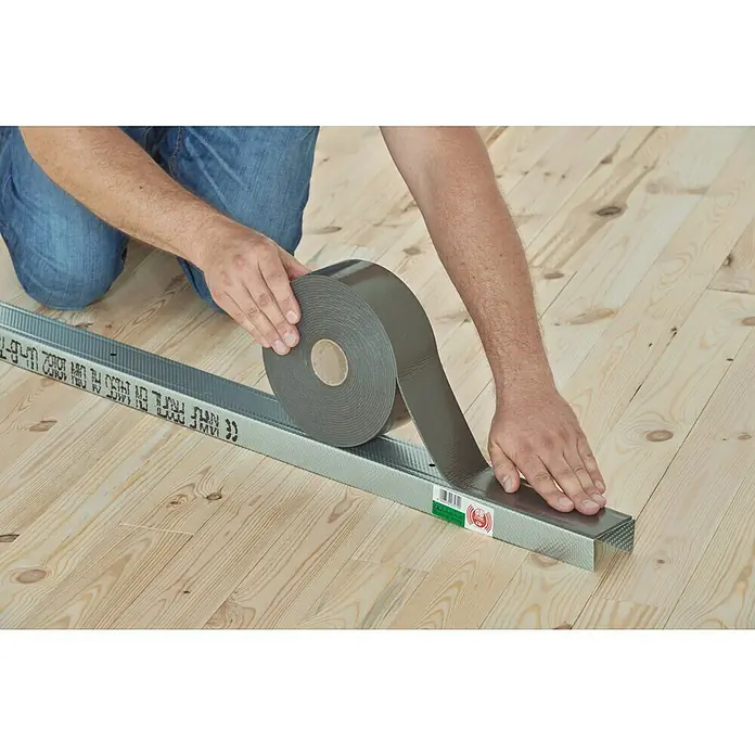 Knauf Dichtungsband - 30 m x 70 mm, Anthrazit - Person befestigt Dichtungsband an einer Metallschiene auf einem Holzboden, um ein abgedichtetes und isoliertes System zu schaffen.