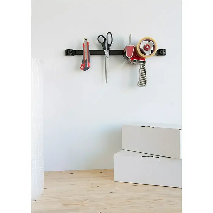 Alpha Tools Magnetleiste - 50 cm, Traglast: 10 kg - An der Wand hängen ein Cuttermesser, eine Schere und ein Klebebandabroller an einer schwarzen Halterung, darunter stehen zwei weiße Kartons.
