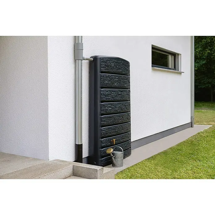 4rain Regenspeicher Extra Slim Stone - 400 l, Grau - Eine dunkle Regentonne mit Wasserhahn und Gießkanne steht neben einem Fallrohr an einer Hauswand. Die Tonne hat eine Steinmuster-Optik.