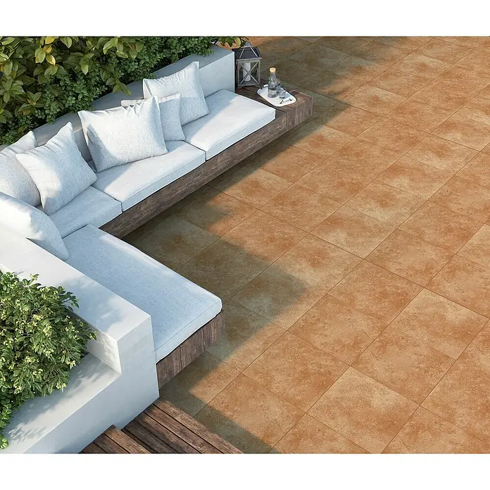 Halcón Pavimento porcelánico Medellín - 33,3 x 33,3 cm, Teja, Mate - La imagen muestra un área exterior con baldosas de color terracota, creando un ambiente cálido. Un sofá elegante con cojines invita a relajarse.