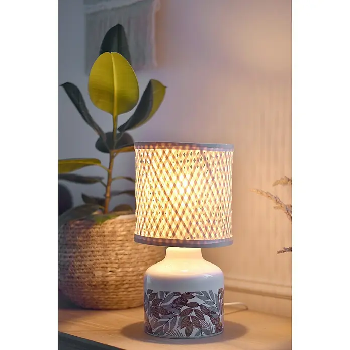 Idp Lampshades Lámpara de sobremesa Penny - 40 W, L x An x Al: 17 x 17 x 32 cm, Marrón, Natural, E14 - Elegante lámpara de mesa con base estampada y pantalla tejida, que emite una luz cálida. Está colocada junto a una planta verde en una cesta.