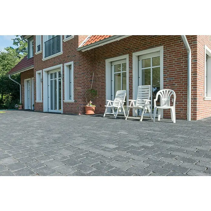Diephaus Pflasterstein AustinBasalt, 28 x 21 x 7 cm, Rechteckig - Außenaufnahme eines gepflasterten Bereichs mit drei weißen Gartenstühlen vor einem Backsteingebäude mit weißen Fensterrahmen.