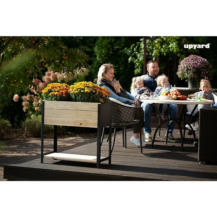 Upyard Hochbeet Modernbox hoch - 78 x 36 x 72 cm, Holz, Natur/Schwarz - Ein erhöhter Pflanzkasten mit gelben Blumen steht auf einer Terrasse, während eine Familie im Hintergrund sitzt und sich entspannt.