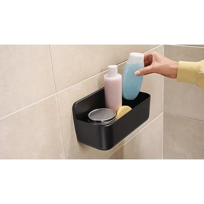 Tesa Cesta de baño Lavaa S - 20 x 11 x 13 cm, Plástico, Negro - Un organizador de ducha negro con productos de baño en una pared de azulejos, una mano colocando una botella azul dentro.
