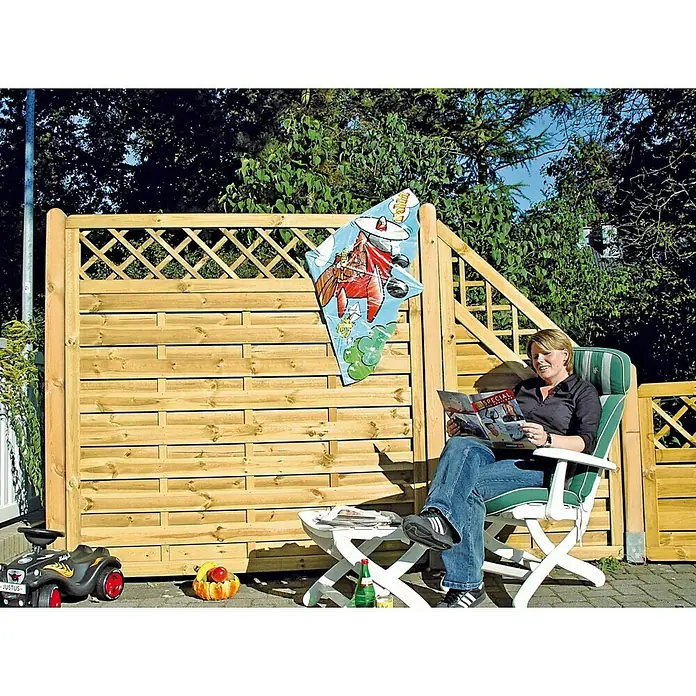 T & J Sichtschutzelement Avantgarde - 180 x 180 cm, Holz, Kesseldruckimprägniert - Frau entspannt im Garten auf einem Stuhl, im Hintergrund ein Holzzaun, der Privatsphäre und natürliche Ästhetik vereint.