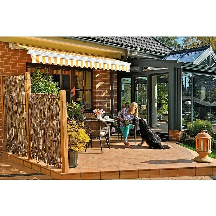 T & J Sichtschutzelement Pepe - B x H: 180 x 180 cm, Holz, Braun - Eine Frau sitzt mit ihrem Hund auf einer Terrasse, die mit einem gelb-weiß gestreiften Vorbau versehen ist und eine gemütliche Atmosphäre schafft.