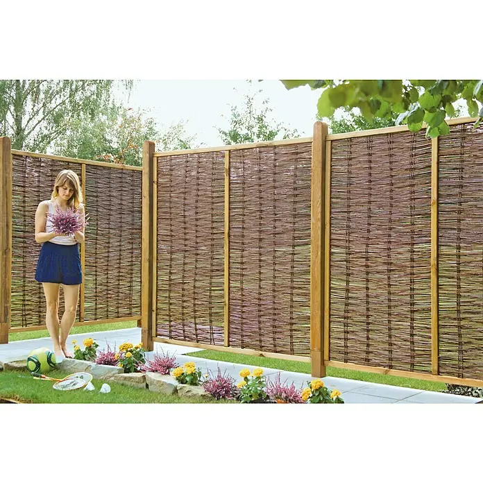 T & J Sichtschutzelement Fogo - 180 x 180 cm, Weide, Braun - Eine Frau steht vor einem stilvollen Sichtschutzzaun aus Weide, der Privatsphäre und natürliche Eleganz bietet. Perfekt für Gärten.