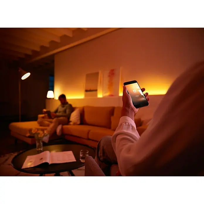 Philips Hue Set de bombillas LED Kit con mando a distancia - E27, Capacidad de atenuación: Intensidad regulable, Blanco cálido, 2.478 lm, 10 W, 3 ud. - Persona usando un smartphone con la app Philips Hue en una sala de estar con iluminación ambiental.