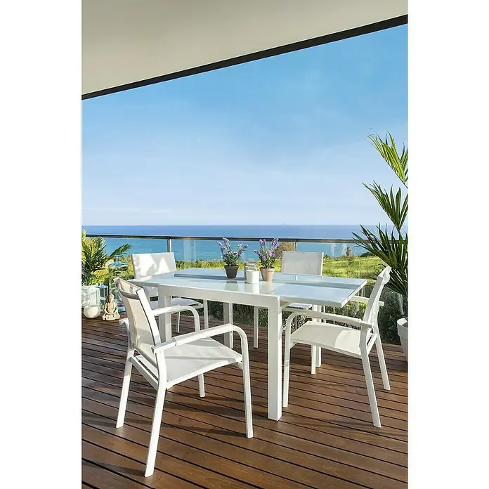 Sunfun Maja Sillón de jardín Maja - An x Pr x Al: 59,5 x 59 x 88 cm, Blanco, Aluminio/Textiline - Una mesa de comedor moderna con sillas blancas en un patio con vistas al mar. El diseño elegante crea un espacio exterior acogedor.