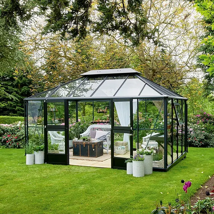 Juliana Gewächshaus Grand Oase - Außenmaß inkl. Dachüberstand (B x T): 439 x 296 cm, Material Seitenwand: Glas, 3 mm, Anthrazit - Ein elegantes Gewächshaus mit schwarzem Rahmen, ausgestattet mit weißen Möbeln und umgeben von üppigem Grün.