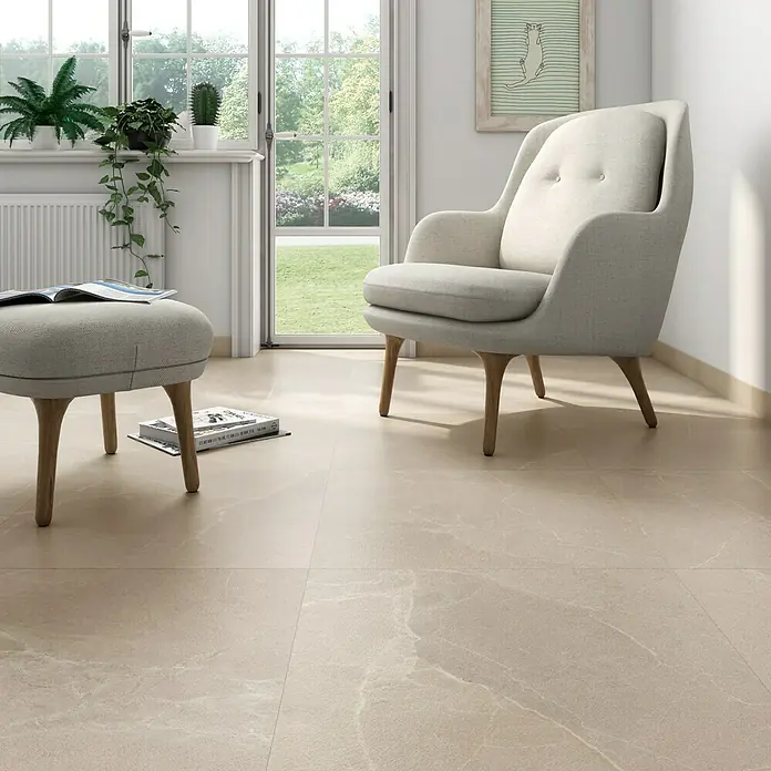 Pavimento porcelánico Soul - 60,8 x 60,8 cm, Base, Mate - Interior acogedor con suelo de baldosas beige, un elegante sillón y un taburete a juego, creando un ambiente armonioso y elegante.
