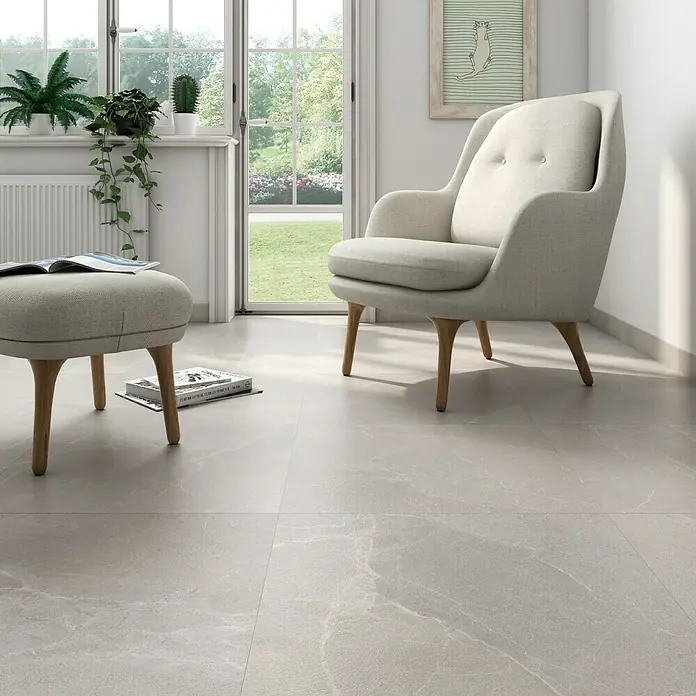 Pavimento porcelánico Soul - 60,8 x 60,8 cm, Light, Mate - La imagen muestra una zona de estar luminosa y moderna con un elegante sillón y un taburete a juego sobre elegantes baldosas grises.
