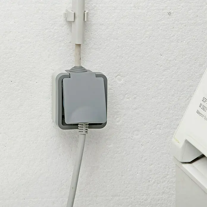 Voltomat Blueline Feuchtraum-Steckdose - 1-fach, Horizontal, IP54, Grau - Graue, wasserdichte Steckdose mit angeschlossenem Kabel an einer weißen Wand.