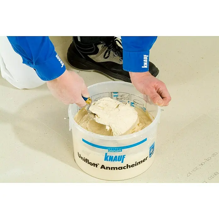 Knauf Fugenspachtel Uniflott - 8 kg - Nahaufnahme von einem Eimer Knauf Uniflott Anmacheimer mit Spachtel, gefüllt mit einer hellen Spachtelmasse, umgeben von Bauarbeitern in Arbeitskleidung.