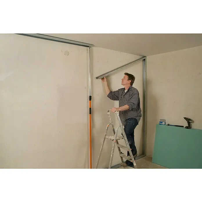 Knauf CD-Deckenprofil - Länge: 2.600 mm, Stahl - Ein Mann auf einer Leiter befestigt eine Metallschiene an einer Wand, um eine neue Wand zu bauen. Werkzeuge und Baumaterialien sind im Raum verteilt.