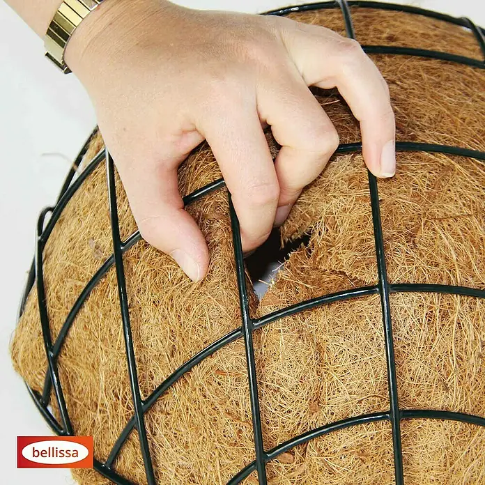 Bellissa Blumenampel Hanging Basket - Ø x H: 35 x 15 cm, Metall, Schwarz - Nahaufnahme einer Hand, die ein Loch in das Kokosfasermaterial eines Bellissa-Pflanzkorbs macht, der von einem schwarzen Drahtgitter gehalten wird.