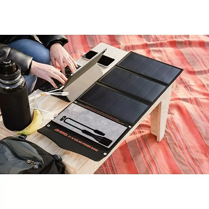Yard Force Cargador solar LX PB21 - 48,1 Wh, 65 x 28,5 x 1,5 cm - Un power bank solar AMIRO 21W desplegado carga una tableta y un teléfono en una superficie de madera, junto a una persona usando la tableta.