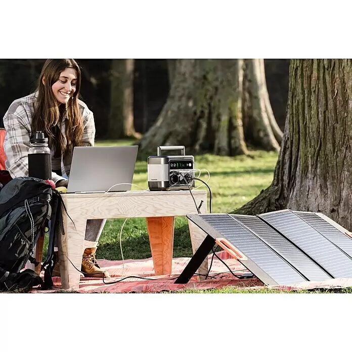 Yard Force Powerstation LX PS300 - 296 Wh, Nennleistung: 300 W, Max. Leistung: 550 W - Frau lädt Laptop im Freien mit schwarzem/silbernem Powerstation und verbundenem dunklem Solarpanel auf.