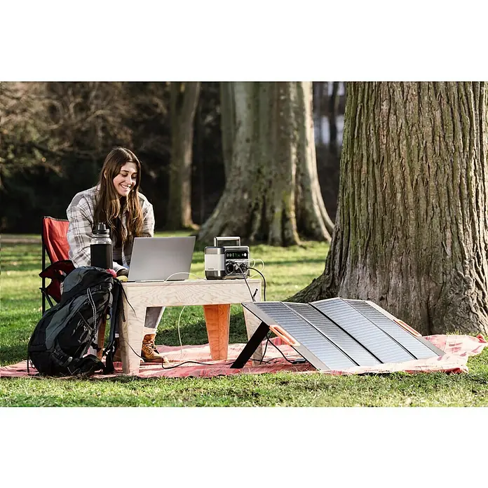 Yard Force Powerstation LX PS300 - 296 Wh, Nennleistung: 300 W, Max. Leistung: 550 W - Eine Frau arbeitet an einem Laptop mit einem tragbaren Solargenerator und Solarpanel im Freien.