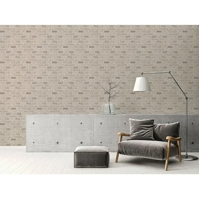 A.S. Création Il Decoro Papiertapete - Taupe, Steinoptik, 10,05 x 0,53 m - Wohnzimmer mit Sessel und Hocker in Grau, ergänzt durch eine Stehlampe und eine Backsteinwand.