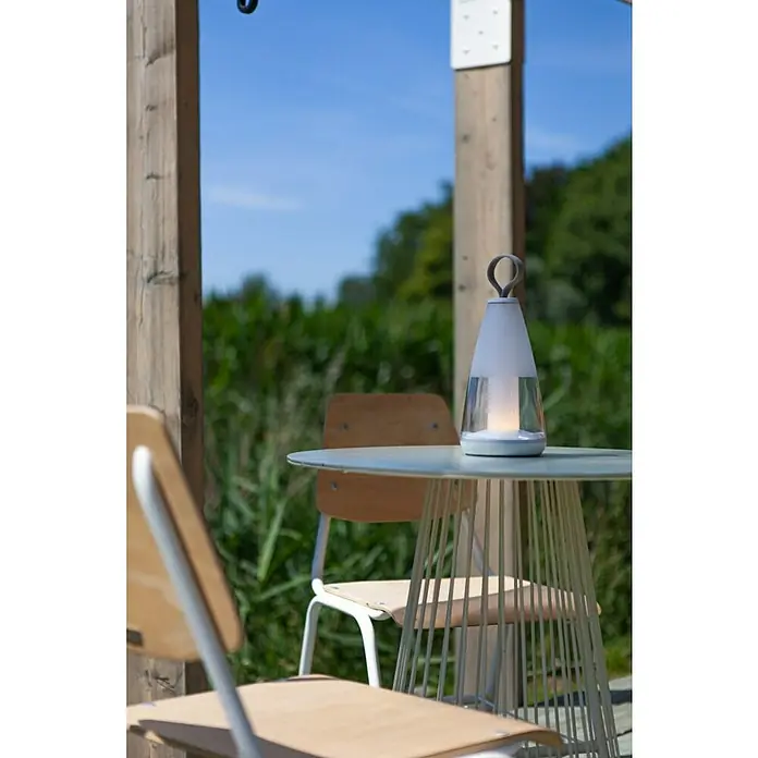 Lutec Akku-Tischleuchte PEPPER - 3 W, L x B x H: 12 x 12 x 29 cm, Weiß, RGBW - Elegante Tischleuchte auf einem Gartentisch, umgeben von Stühlen und üppigem Grün. Die Leuchte spendet warmes Licht und schafft eine angenehme Atmosphäre.