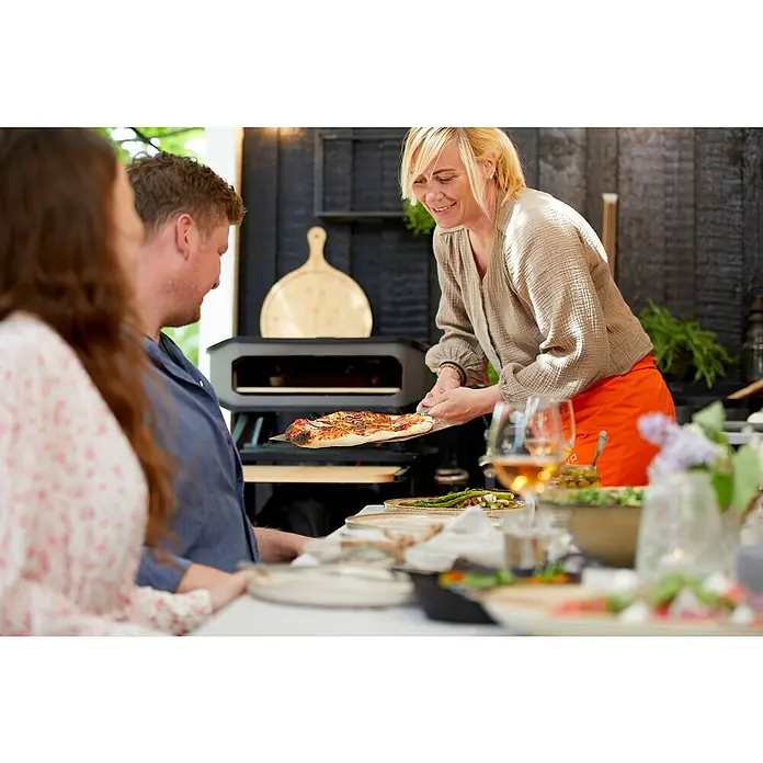 Cozze Horno para pizza gas 17" - 8 kW - Una mujer sirve una pizza recién hecha de un horno de pizza portátil al aire libre a sus invitados.