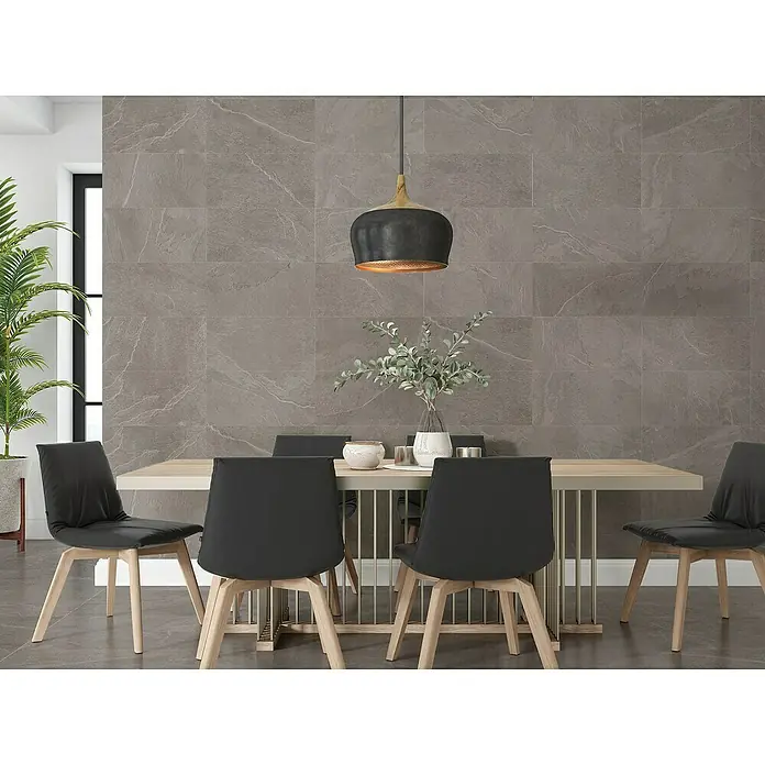 Halcón Pavimento porcelánico Aura - 30 x 60 cm, Ceniza, Mate, Espesor: 8 mm - Un primer plano de un moderno juego de comedor con mesa, sillas e iluminación colgante contra una pared de piedra gris.