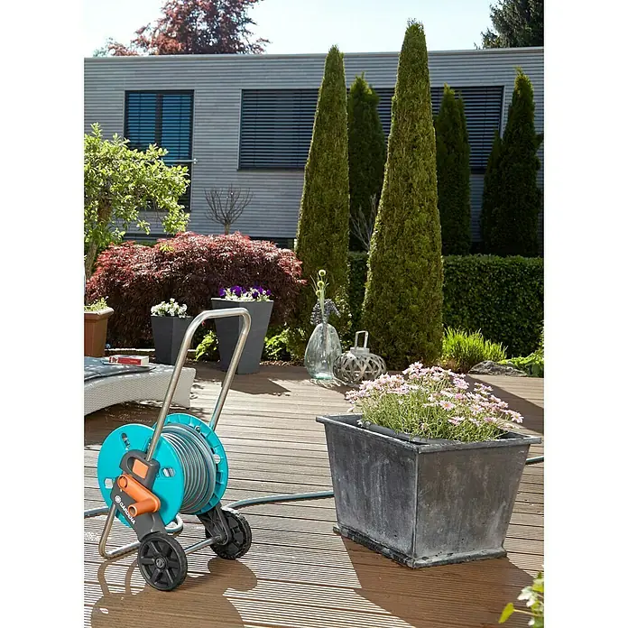 Gardena Schlauchwagen-Set CleverRoll S - Kunststoff/Metall, Schlauchlänge: 20 m - Eine Gartenszene mit einem Gardena-Schlauchwagen im Vordergrund, der auf einer Holzterrasse steht, mit Pflanzen und einem modernen Haus im Hintergrund.