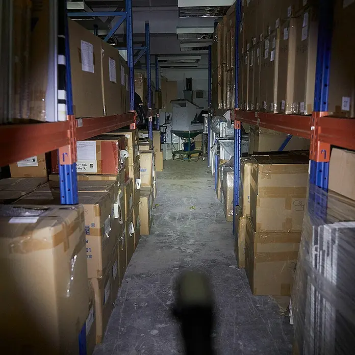 GP Taschenlampe C34 - 85 lm - 550 lm, IPX4 - Blick in ein Lagerhaus mit Regalen voller Kartons, beleuchtet von einem Lichtstrahl, der die Waren hervorhebt. Die Lagerlogistik wird durch die Gänge betont.