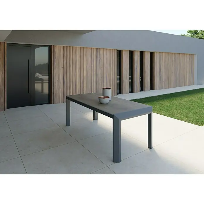 Mesa de jardín Mistral - L x An x Al: 192 x 102 x 74 cm, Aluminio/Cerámica, Antracita - Una mesa gris con dos elementos decorativos se sitúa en un patio frente a una casa moderna con pared de madera.