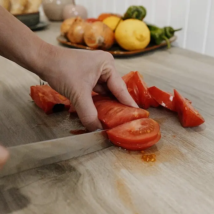 Mesa de jardín Mistral - L x An x Al: 192 x 102 x 74 cm, Aluminio/Cerámica, Antracita - Primer plano de una mano usando un cuchillo para cortar un tomate rojo sobre una mesa de madera, con otros ingredientes al fondo.