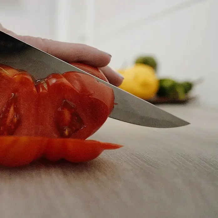 Mesa de jardín Mistral - L x An x Al: 192 x 102 x 74 cm, Aluminio/Cerámica, Antracita - Vista de cerca de una persona cortando un tomate rojo jugoso sobre una mesa de madera con un cuchillo afilado, enfatizando ingredientes frescos.