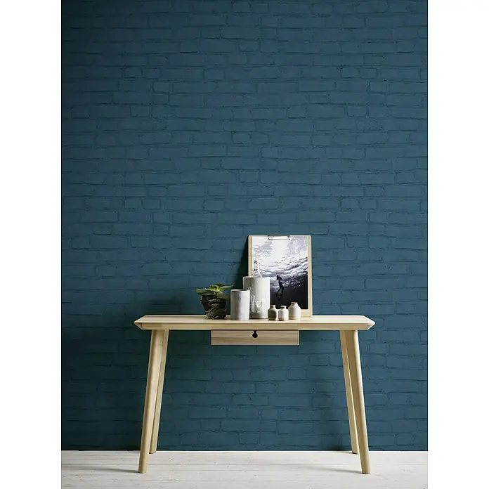 A.S. Création Neue Bude 2.0 ED II Vliestapete Steine - Dunkelblau, Steinoptik, 10,05 x 0,53 m - Elegantes Interieur mit einem hellen Holztisch, der mit dekorativen Elementen und einem gerahmten Bild verziert ist, vor einer stilvollen Backsteinwand.