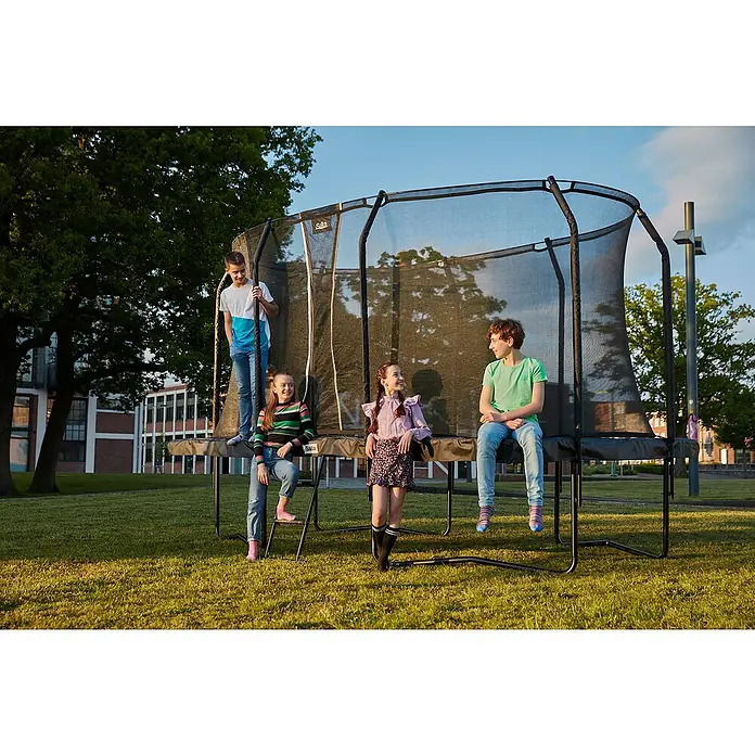 Salta Trampolin Premium Black Edition - 396 x 244 cm, Schwarz - Eine Gruppe Kinder sitzt und steht auf einem Salta-Trampolin im Freien, umgeben von Bäumen und Gebäuden.